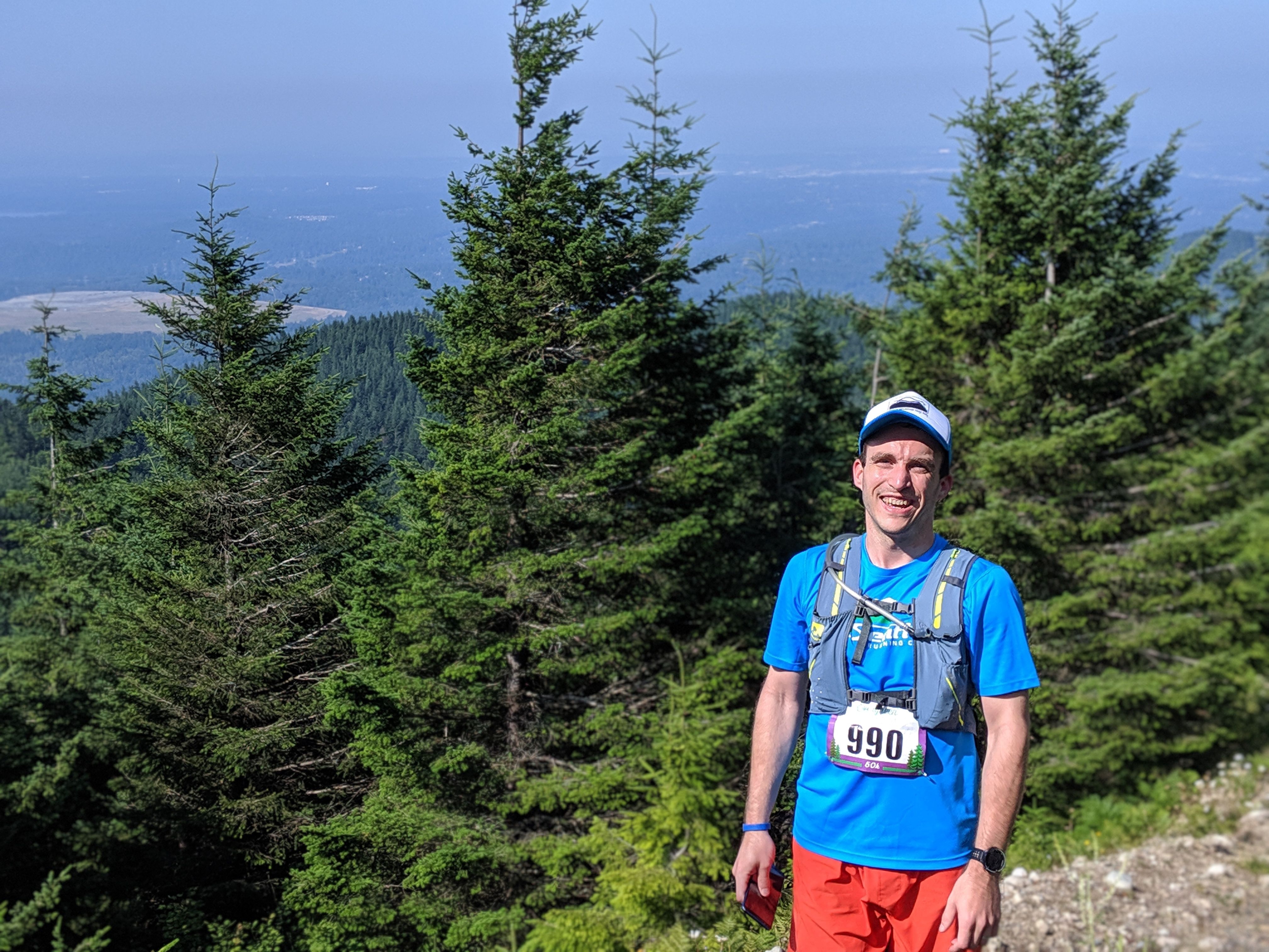 Photo of Bobby at the top of a hill, with some coniferous trees behind him, and in the distance rolling hills and a lake