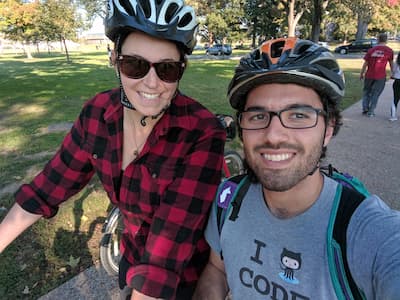 A selfie of me and Kristen in bike helmets in a city park. Kristen is in a red checkered flannel button-up shirt, and I'm in a tshirt that says “I love code”, where the word “love” is replaced by the GitHub octocat logo