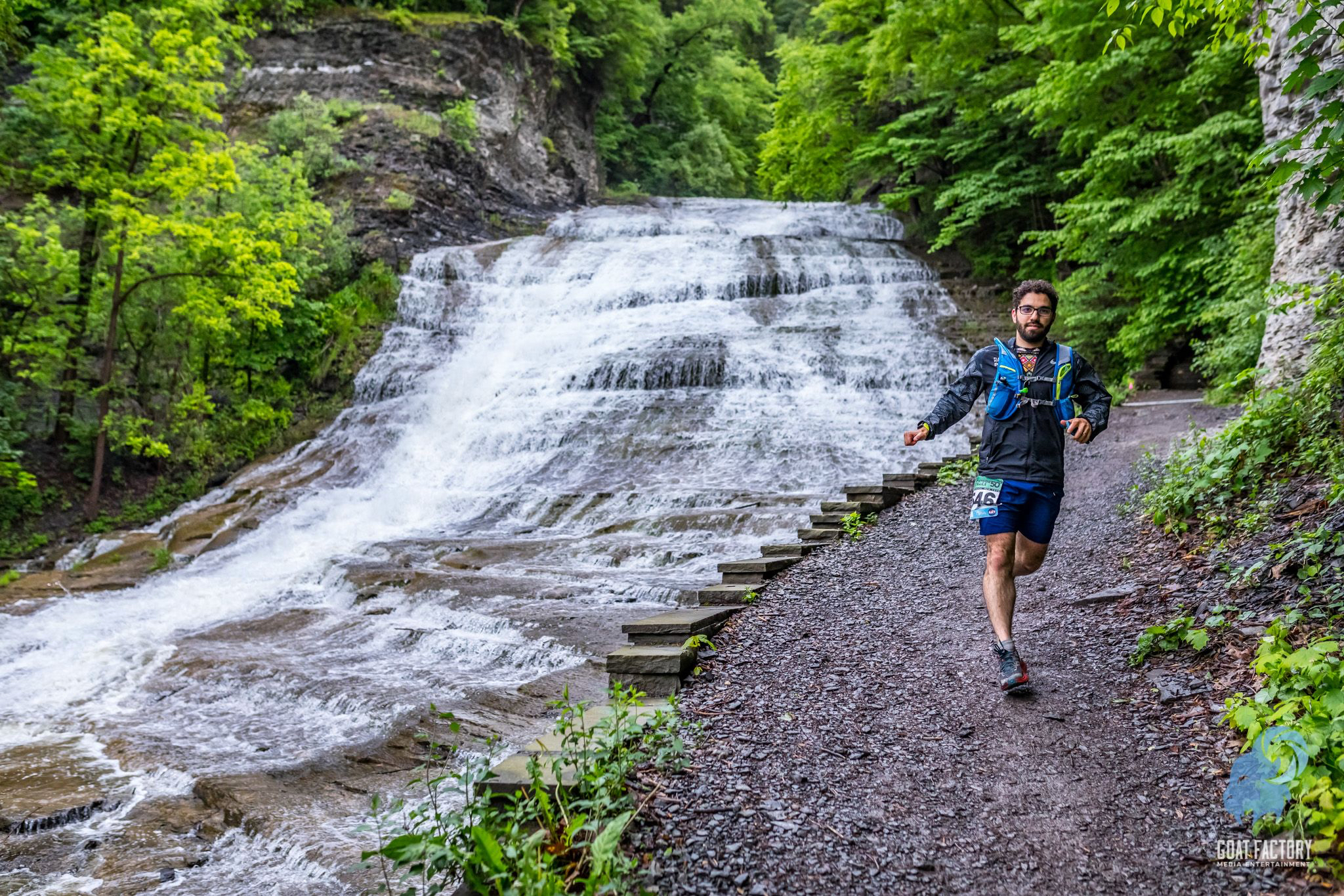 Me running down a gravel path with a waterfall to the left. I'm wearing a black windbreaker, a blue hydration pack, shorts, and a race bib. I have a relaxed yet focused look on my face.