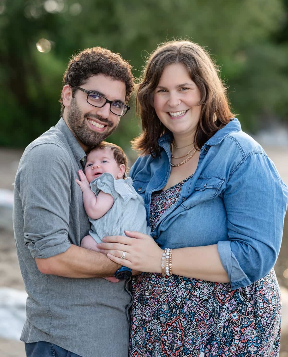 Kristen and Josh, embracing and smiling at the camera. Josh is holding a newborn baby in a gray gress. Josh is in a gray button-up shirt, and Kristen is wearing a denim shirt over a floral dress.