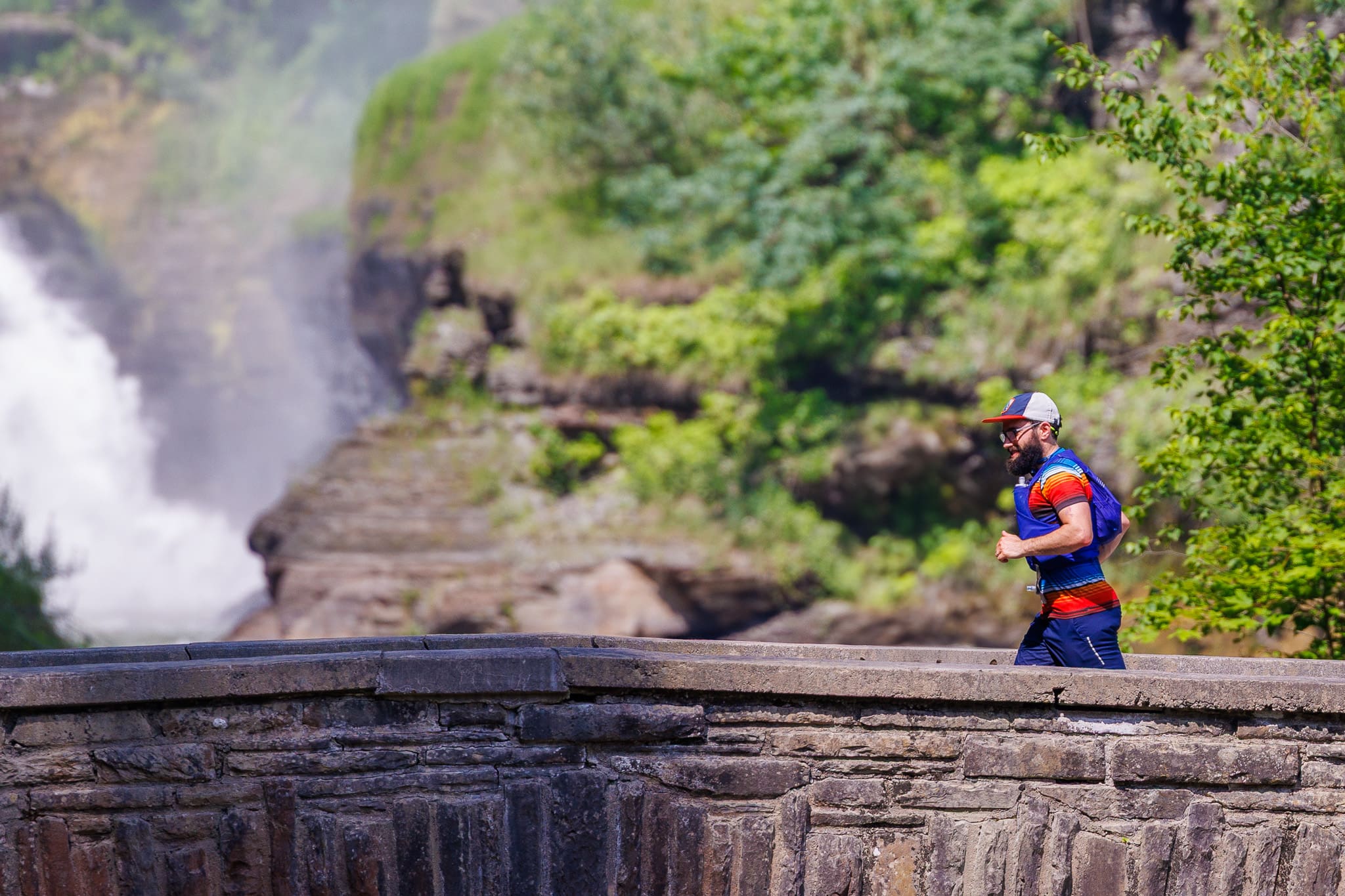 Josh, with a thick black beard, running across a stone bridge with a blurry waterfall in the background. Josh is dressed in a colorful running shirt, a blue hydration vest, and a brimmed running hat.
