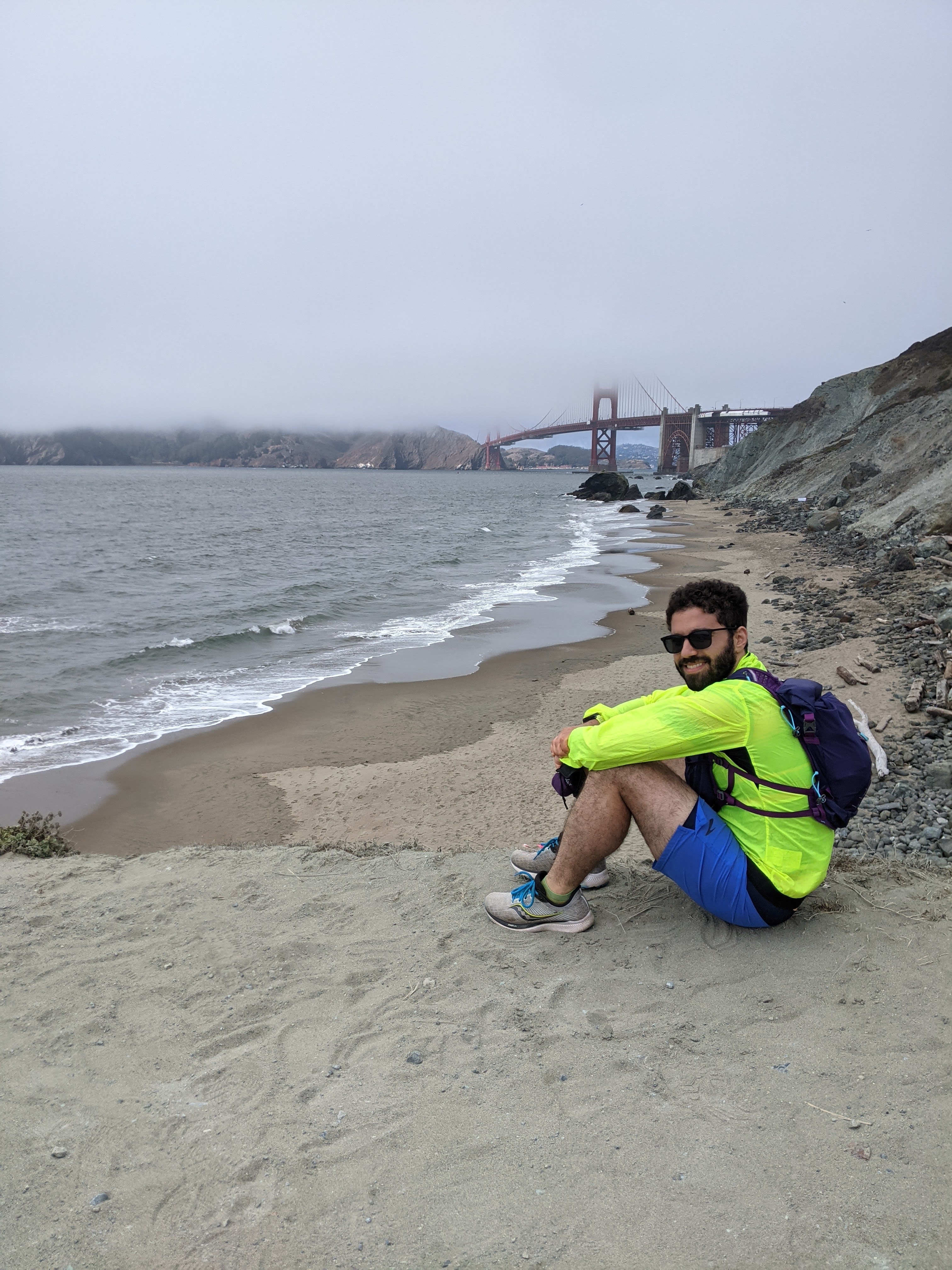 Me in a neon yellow windbreaker, sunglasses, and a hydration vest, sitting on a beach. In the distance is the Golden Gate Bridge, shrouded in fog.